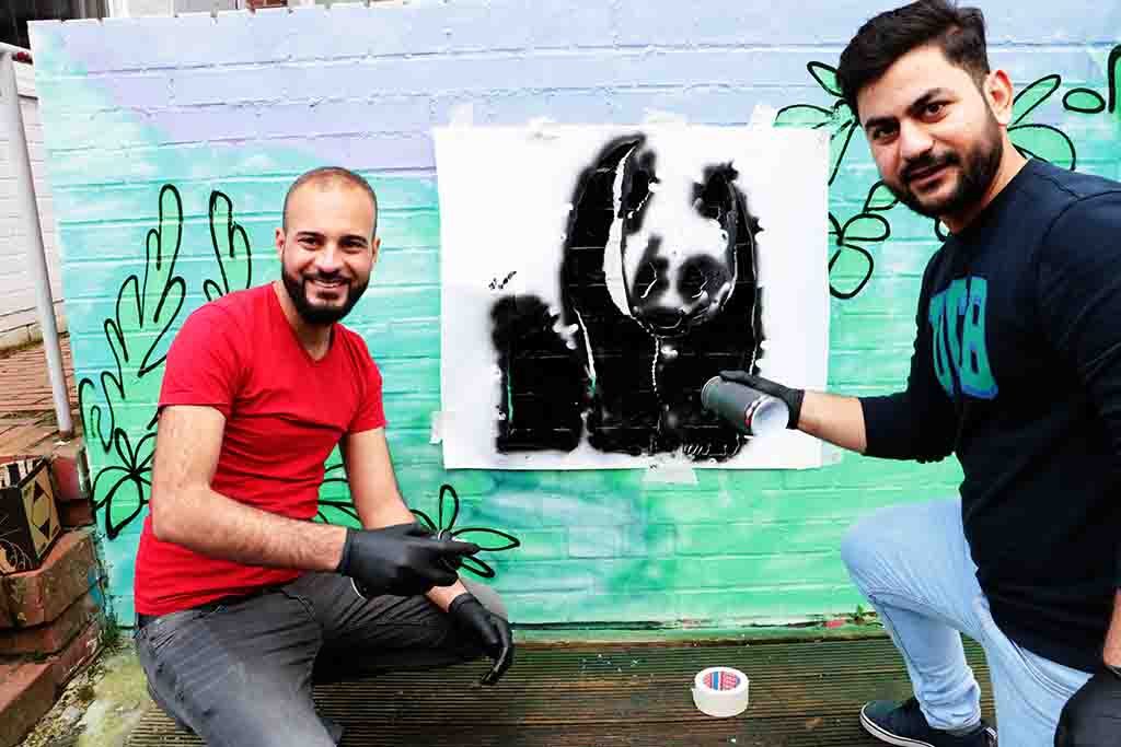 Zwei Männer tragen mit einer Schablone das Bild eines Pandabären auf eine Wand auf.